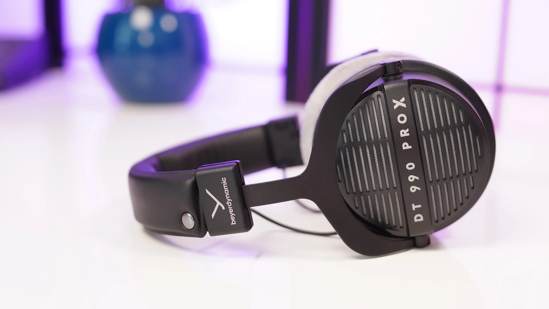 The Beyerdynamic DT 990 PRO X headphones are shown in a side profile, resting on a white surface with a blurred background featuring purple lighting and a decorative object.