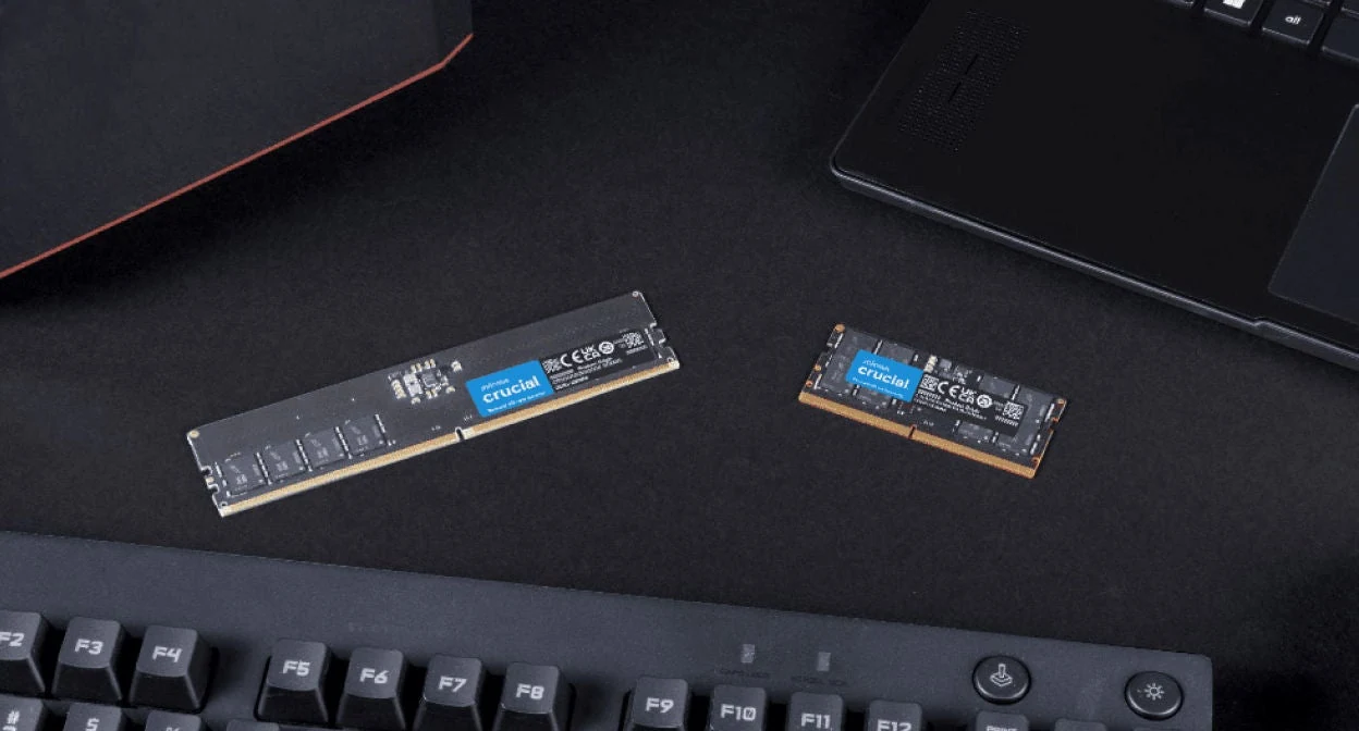 The image shows two Crucial memory modules placed on a desk with part of a keyboard and laptop visible in the foreground.