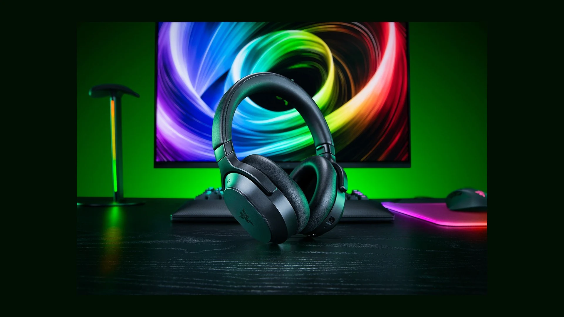 A black Razer Barracuda V2 Pro headset is displayed on a desk with a colorful background featuring a monitor and other gaming peripherals.