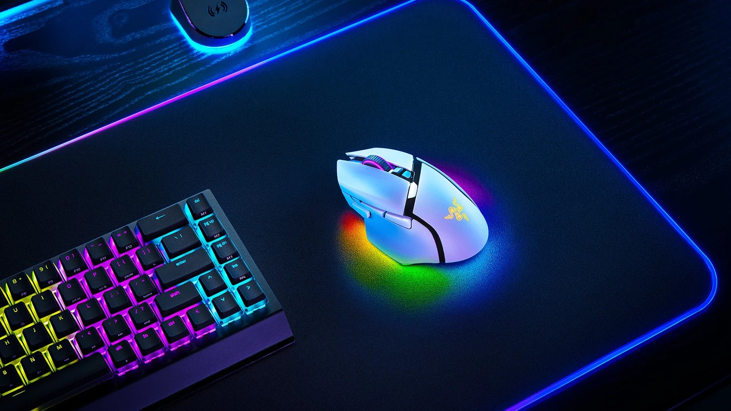 A white Razer mouse with a yellow logo is placed on a black mousepad with blue lighting, next to a keyboard with rainbow-colored backlighting.