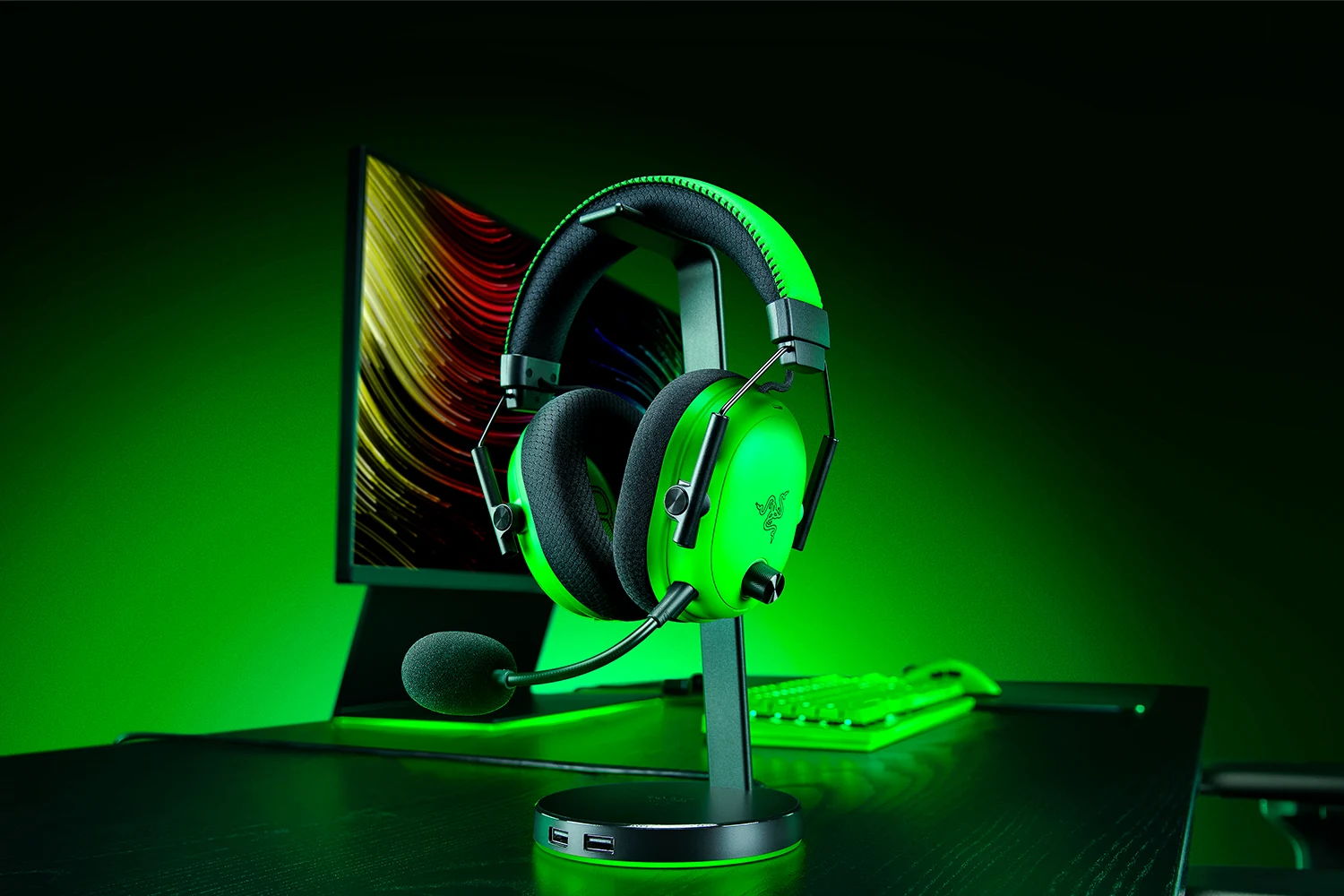 A green Razer BlackShark V3 Pro Esports headset is displayed on a stand, with its microphone extended and positioned in front of a monitor displaying colorful graphics.