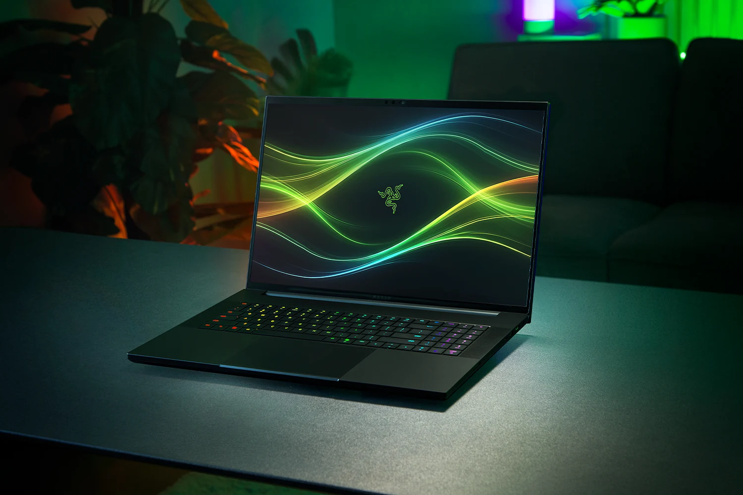 A Razer Blade 18 gaming laptop with a black finish, positioned on a table in a room illuminated by green and orange ambient lighting.