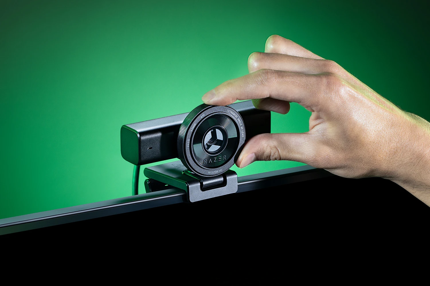 A hand holding a Razer Kiyo V2 webcam, showing the front lens and part of the body against a green background.