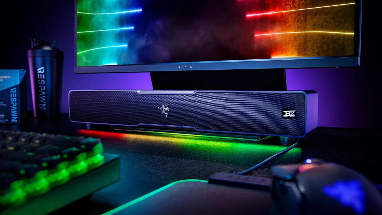A black Razer soundbar with a textured front and illuminated RGB lighting is placed on a desk, surrounded by other gaming peripherals.