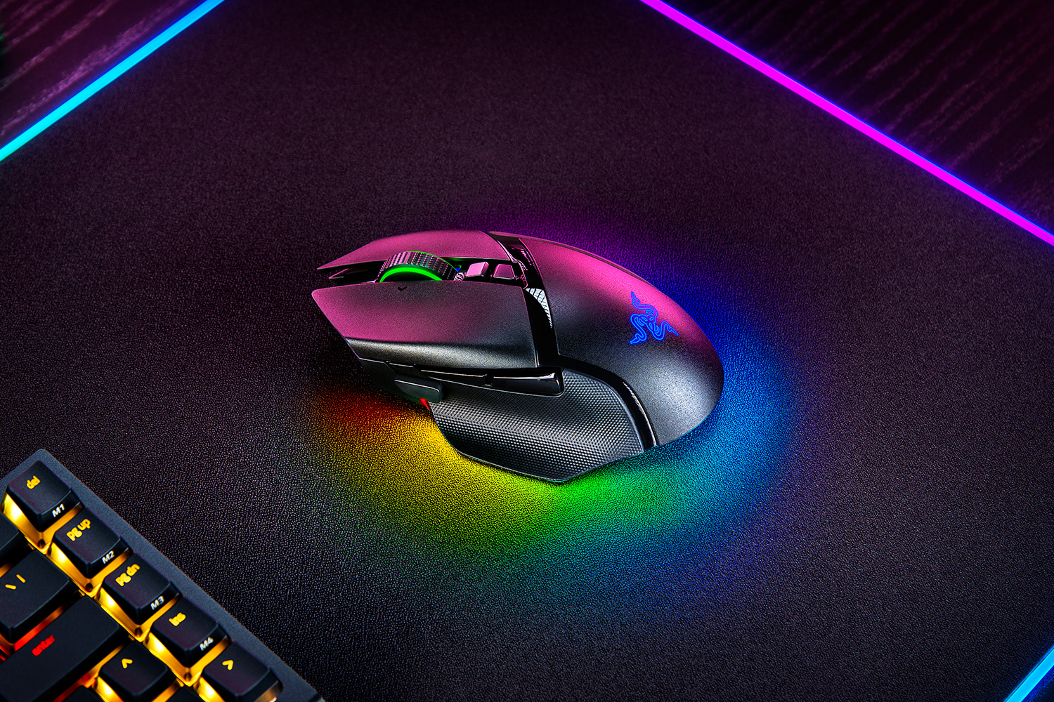 A Razer Basilisk V3 Pro mouse is shown in a top-down view with its RGB lighting illuminated, placed on a textured surface next to a keyboard.