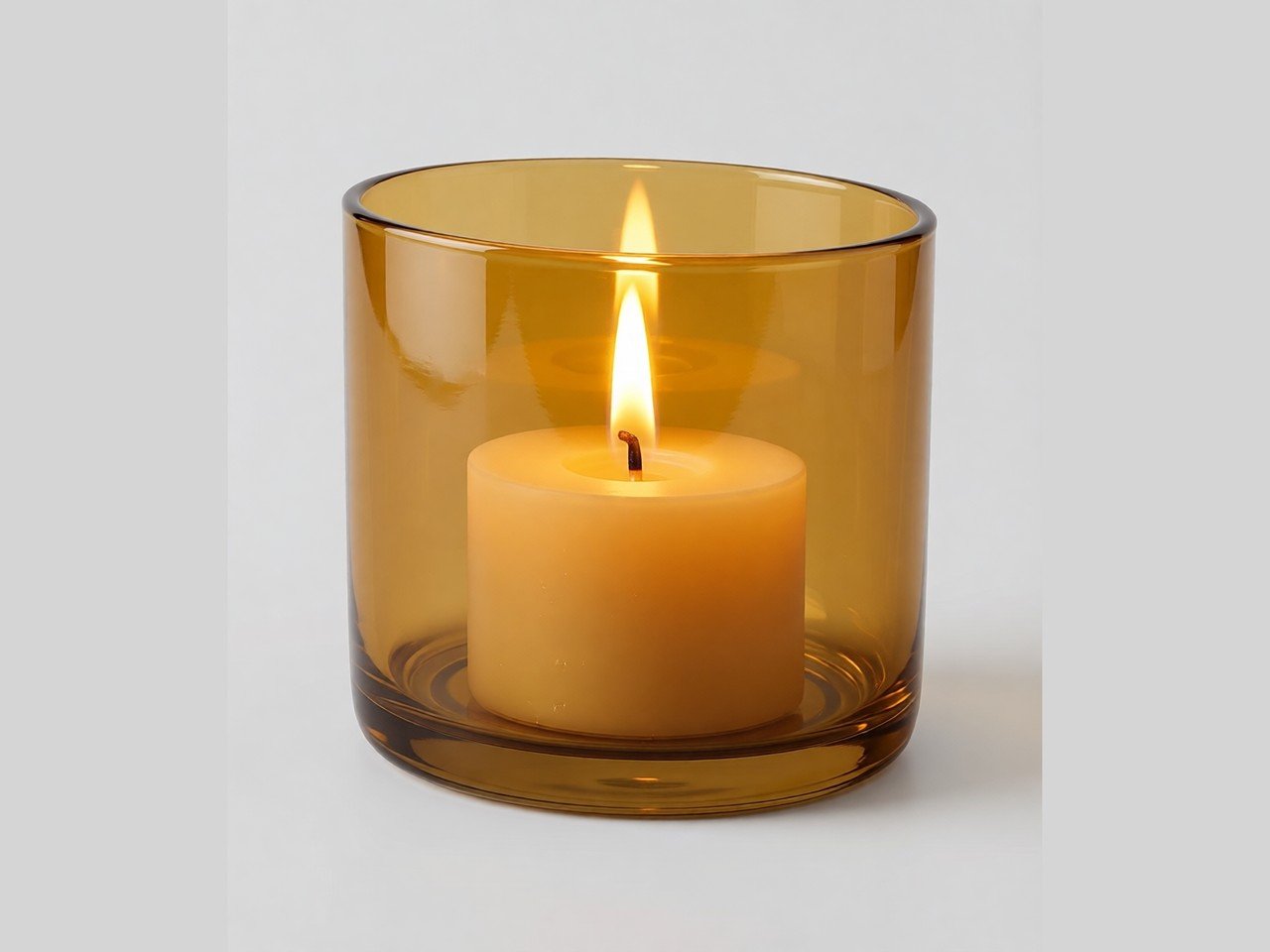 A cylindrical amber glass candle holder containing a lit yellow candle, on a plain grey background.