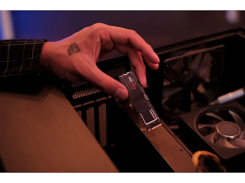A hand holding a Samsung 990 PRO SSD in front of an open computer case.