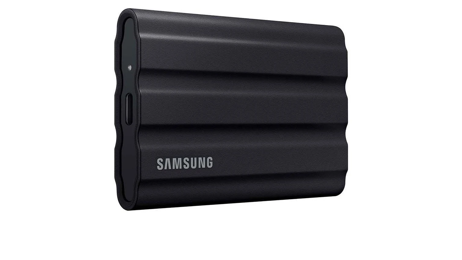 A black Samsung T7 Shield portable SSD shown at a three-quarter angle on a white background, with its ribbed rubber exterior, USB-C port and Samsung wordmark visible.